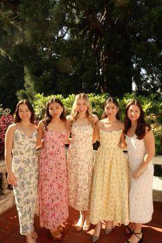 four women in dresses standing next to each other on a brick floored area with trees and bushes behind them