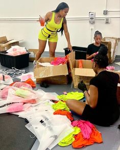 two women in yellow shirts and black shorts sitting on the floor next to boxes filled with clothes