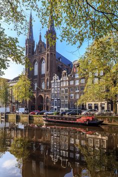 an old church is reflected in the water near some trees and boats on the river