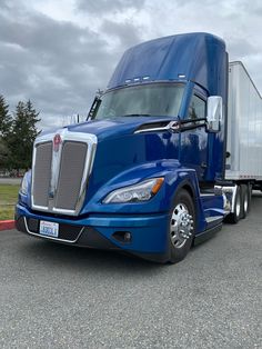 a blue semi truck is parked in a parking lot with trees and grass behind it