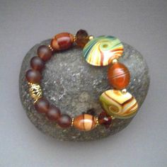 The beautiful 7-inch Southwestern-Boho-themed Mixed Stretch Bracelet of Porcelain Disks with vivid bands of Green and Dark Orange and Lampwork Glass beads is perfect for layering with other gold and gemstone bracelets.  An exquisite 16 X 12mm Handmade Orange with Gold-banded Lampwork Glass Bead is the central bead with Two large 25 X 20mm Polished Porcelain Disks with Green, Orange and Tan veins on either side.  Faceted 11 X 9 Czech Glass Rondel Beads in a Clear Sparkly Brown lead into 16 X 12mm Bohemian Round Brown Beads, Bohemian Czech Glass Beads With Large Beads, Bohemian Brown Beaded Bracelets With Oval Beads, Bohemian Czech Glass Oval Beads, Bohemian Czech Glass Large Beads, Bohemian Oval Czech Glass Beads, Handmade Brown Czech Glass Beads, Bohemian Czech Glass Polished Beads Bracelets, Bohemian Czech Glass Orange Beaded Bracelets