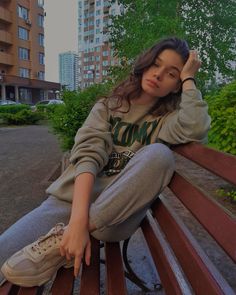 a young woman sitting on top of a wooden bench in front of tall apartment buildings