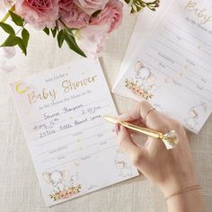 a person writing on a baby shower card next to pink flowers and a gold pen