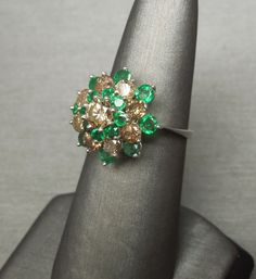 "Vintage Mid-Century Circa 1960 Constructed of 18KT White Gold Containing 9 Genuine Earth-Mined Round Brilliant cut Natural Fancy Cognac Diamonds weighing a total of approximately 1.65 carats Center Diamond - VS2-SI1 Clarity Surrounding Diamonds - I1-I2 Clarity with 16 Genuine Earth-Mined Round cut Natural Intense Green Emeralds weighing a total of approximately 1.20 carats Cluster measuring approximately 0.70\" & top section of ring measures about 11.2mm high when on the finger Inside shank Formal Cluster Emerald Ring, Formal Multi-stone Cluster Diamond Ring, Vintage Green Cluster Ring With Halo Setting, Vintage Cluster Emerald Ring For Anniversary, Classic Multi-stone Cluster Ring For Formal Occasions, Classic Multi-stone Cluster Ring For Formal Events, Green Multi-stone Diamond Ring For Formal Occasions, Formal Green Multi-stone Diamond Ring, Vintage Green Diamond Cluster Ring