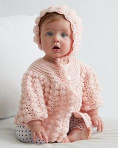 a baby wearing a pink crochet sweater and bonnet sits on a bed with pillows