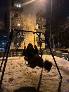 a swing set in the middle of a snowy yard at night with buildings in the background