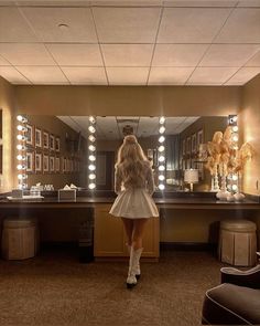 a woman standing in front of a mirror with lights on the wall and vanity behind her