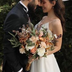 a bride and groom standing together in front of some trees with their wedding bouquets