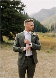 a man wearing a suit and tie standing in the middle of a field with mountains behind him