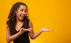 a woman with curly hair is holding out her hands and smiling while standing against a yellow background