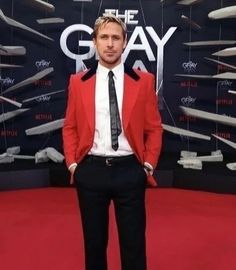 a man in a red jacket and tie standing on a red carpet with shoes hanging from the ceiling