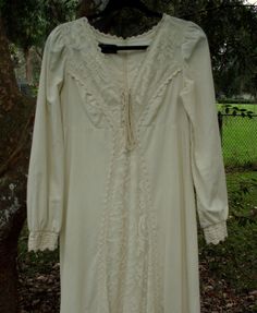 "Today I have for you a lovely 1969 Romance/Ren style Gunne Sax dress of medium to heavyweight, raw cotton that is accented with floral and heavyweight, crocheted lace. This design was often used as a wedding dress in that era and would suffice as one today. The condition is excellent and the craftsmanship is superb. It all starts with the tie bodice that is covered with a lace that consists of individual flowers within a free formation (also to be found on the attached apron). Heavy, durable la Cream Cotton Dress With Crochet Trim, Vintage Cotton Dresses With Lace Patchwork, Bohemian Cotton Dress With Lace Collar, Vintage Cotton Dress With Lace Cuffs, Fitted Cotton Lace Dress With Crochet Trim, Fitted Dress With Crochet Trim In Cotton Lace, Long Sleeve Cotton Dress With Lace Cuffs, Fitted Cotton Dress With Lace Work, Long Sleeve Lace Dress With Crochet Trim