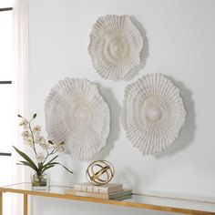 two white wall hangings on the side of a table with flowers in vase and books