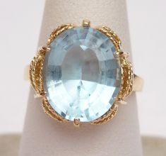 This is a classic birthstone ring from the 1960s. This ring has an oval step cut synthetic blue stone in the center that measures 12 x 10 mm. The cut is very different than a normal oval cut stone, so it has a neat look to it. The stone is set in 6 prongs and the ring has a cut out scalloped design around the stone. The inside of the ring is stamped JUB 100 10 (the K has worn off) and the ring has been tested to verify that it is 10 karat yellow gold. The ring weighs 3.9 grams and is a size 5.75 Oval Topaz Ring For Formal Occasions, Vintage Oval Topaz Birthstone Ring, Vintage Oval Blue Topaz Rings, Formal Oval Cabochon Topaz Ring, Vintage Oval Topaz Gemstone Ring, Vintage Blue Oval Ring, Vintage Oval Rings With Large Stone, Scalloped Design, Step Cut