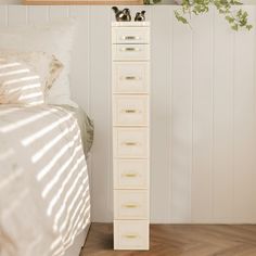 a tall white drawer sitting on top of a wooden floor next to a bed and plant