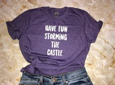 "A fun, and soft shirt for fans of The Princess Bride. This comfy shirt makes an awesome gift and goes well with everything from jeans and boots to leggings and sneakers. *Processing time is currently 4-9 business days. Thank you for your patience!* Design: \"Have Fun Storming The Castle\" Shirt Sizing Softstyle (Unisex Adult T-Shirt) - Comfortable for both women and men to wear. - Please see size chart in the photos for more details. Materials - 100% preshrunk ringspun cotton - Heather colors 3 Casual Purple T-shirt For Fan Merchandise, Fun Slogan T-shirt For Fan Merchandise, Casual Purple T-shirt For Fans, Casual Pre-shrunk Shirt For Fan Conventions, Casual Tops With Funny Text For Gifts, Unisex Casual Shirt With Funny Text, Casual Shirt With Funny Text As Gift, Casual Purple Slogan T-shirt, Casual Purple T-shirt As Gift