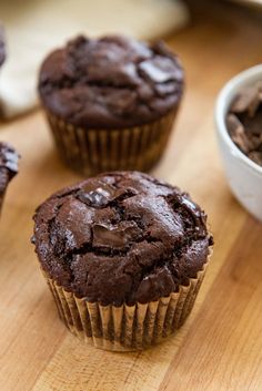 three chocolate muffins sitting on top of a wooden table next to a bowl of chocolate chips