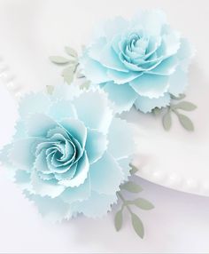 two blue paper flowers sitting on top of a white cake plate next to each other