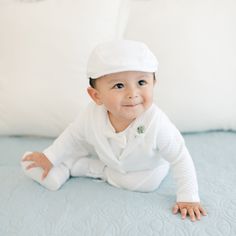 A baby boy in the Elijah 3-Piece Suit and cap is sitting on a light blue blanket. The outfit, perfect for a special occasion, features textured cotton. He has a slight smile and is looking slightly upward, with white pillows in the background. Classic White Sets For Baptism, Baby Boy Formal Wear, Christening Dress For Baby Boy, Baby Boy Suit Formal, Handsome Outfit, Traditional White Baptism Sets, Christening Clothes For Baby Boy, Baby Boy Christening Outfit, Boy Dress