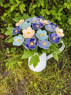 a white pitcher filled with blue flowers on top of a grass covered field next to bushes