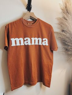 "Introducing the Mama T-shirt, exclusively available on Powered by Daisies! Celebrate motherhood in style with this comfortable and chic rust orange t-shirt. Made from high-quality materials, this t-shirt features the word \"Mama\" in bold letters. It's the perfect gift for yourself or any mom in your life - whether you're a new mom, a seasoned pro, or somewhere in between. Available in a range of sizes, our Mama T-shirt is designed to fit all body types comfortably. It's perfect for running err Boho Mama Shirt, Brown Short Sleeve T-shirt With Letter Print, Brown Text Print Crew Neck T-shirt, Brown Crew Neck T-shirt With Text Print, Pre-shrunk Brown Crew Neck T-shirt, Brown Cotton T-shirt With Letter Print, Brown Relaxed Fit Pre-shrunk T-shirt, Orange Short Sleeve T-shirt With Letter Print, Family Matching Cotton T-shirt Gift