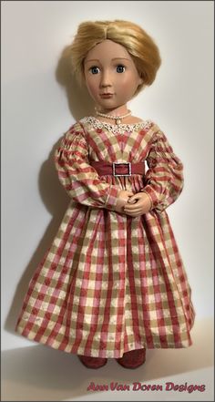 a doll with blonde hair wearing a red and white checkered dress, standing in front of a wall