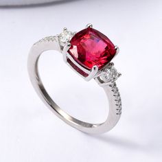 Ruby Engagement Ring Our Ruby Gemstone Ring is crafted With rhodium over 925 Sterling Silver. The Engagement ring features a cushion-cut lab-grown ruby measuring 8x8mm as the centerpiece. It is complemented by two 3mm round moissanite stones and fourteen 1.0mm round moissanite stones. Weighing 2.73 grams, this Silver Ruby Ring is perfect for July birthstone jewelry. It's a thoughtful gift for women who love ruby and gemstone rings. Ruby Ring For Her The red ruby is known for its symbolic meaning Red Cushion Cut Ring With Accent Stones, Three Stone Cushion Cut Ring, Anniversary Lab-created Ruby Cushion Cut Jewelry, White Gold Cushion Cut Ruby Ring With Prong Setting, Silver Three-stone Ruby Ring, Elegant Silver Ruby Ring With Cushion Cut, Elegant Three Stone Lab-created Ruby Jewelry, Elegant Silver Ruby Three Stone Ring, Silver Ruby Ring With Cushion Cut For Anniversary