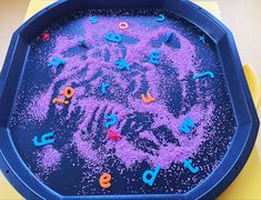 a child's play tray with letters and numbers in purple sand on the table