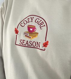 the cozy girl season logo is embroidered on a white sweatshirt with autumn leaves around it