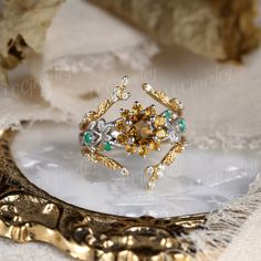 a gold ring with green and white stones on it sitting on a table next to some leaves