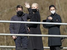 two people in black coats and masks are standing on a bridge with their hands up