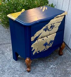 a blue and gold painted dresser sitting on the side of a sidewalk next to a bush