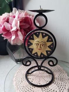 a decorative candle holder with a sun face on it and a pink flower in the background