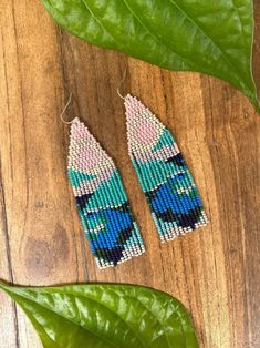 the beaded earrings are sitting on top of a wooden table next to a green leaf