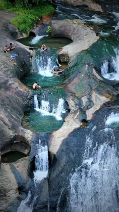 people are swimming in the water near some rocks and waterfall cascading, while another person is laying on his stomach