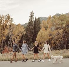 four women walking down the road holding hands with a dog in front of some trees