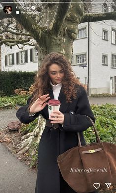 a woman holding a coffee cup in her right hand while standing next to a tree