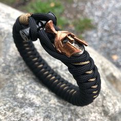 The Undead Spartan features a handmade limited combined copper/white brass bead on our signature weave and closure. Spiced up with additional microcord stitching.  Default colors are black and gold/tan stitch, but any colors can be done on your choice. Gold Braided Bracelet With Adjustable Nylon Cord, Handmade Black Metal Braided Bracelets, Handmade Metal Braided Bracelets In Black, Handmade Black Jewelry With Nylon Cord, Handmade Black Nylon Cord Jewelry, Handmade Gold Braided Bracelet, Handmade Gold Braided Bracelet With Nylon Cord, Adjustable Braided Brown Jewelry, Adjustable Brown Braided Jewelry