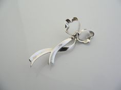 Overview:Offered for sale is a wonderful and very pretty Tiffany and Co. Sterling Silver Ribbon Brooch / Pin. The piece is made from bright and substantial Tiffany silver fashioned to look like a flowing bow / ribbon - very striking and pretty! In excellent condition, this piece is one that you will turn to very often to jazz up your wardrobe. It is no longer produced, and thus makes a great addition for any Tiffany collector. The piece has a perfectly working locking mechanism, and is fully hal Elegant Silver Heart Brooches, Elegant Silver Heart-shaped Brooches, Elegant Silver Heart Brooch, Elegant Heart-shaped Wedding Brooch, Elegant Silver Heart-shaped Brooch, Elegant Silver Brooch With Bow, Silver Wedding Brooch With Ribbon, Formal Silver Brooch With Ribbon, Elegant Brooches With Decorative Bow For Anniversary