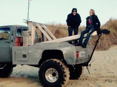 two people sitting on the back of a pick up truck in the sand with an american flag flying above them