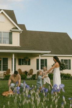 a woman and two children playing in front of a house with purple flowers on the lawn