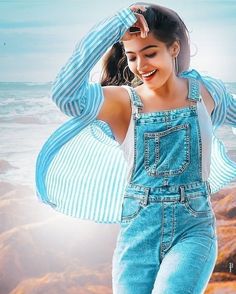 a woman in overalls standing on the beach with her arms behind her head and smiling