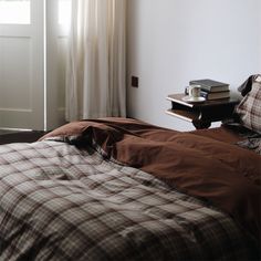 an unmade bed with brown and white plaid comforter on it next to a window