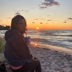 a woman sitting on top of a sandy beach next to the ocean at sunset with a cup of coffee in her hand