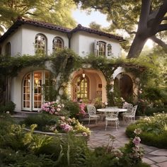 a white house surrounded by flowers and greenery with an outdoor dining table in the foreground