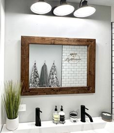 a bathroom mirror sitting above a white sink under three light fixture lights and a potted plant in front of it