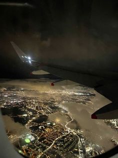 an airplane wing flying over a city at night with lights on the buildings and clouds