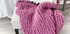 a pink knitted blanket sitting on top of a chair next to a white pillow