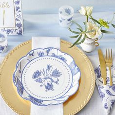 a blue and white place setting with flowers in vases, napkins and gold rimmed plates