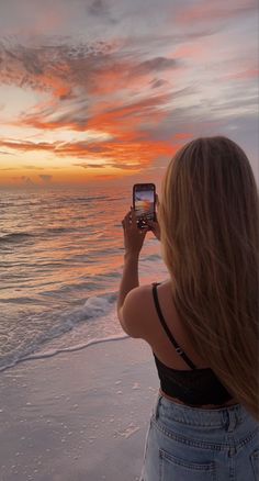 a woman standing on the beach taking a photo with her cell phone at sunset or dawn